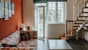 A spacious living room with a high ceiling, a wooden staircase, and a large window with a view of the outdoors.