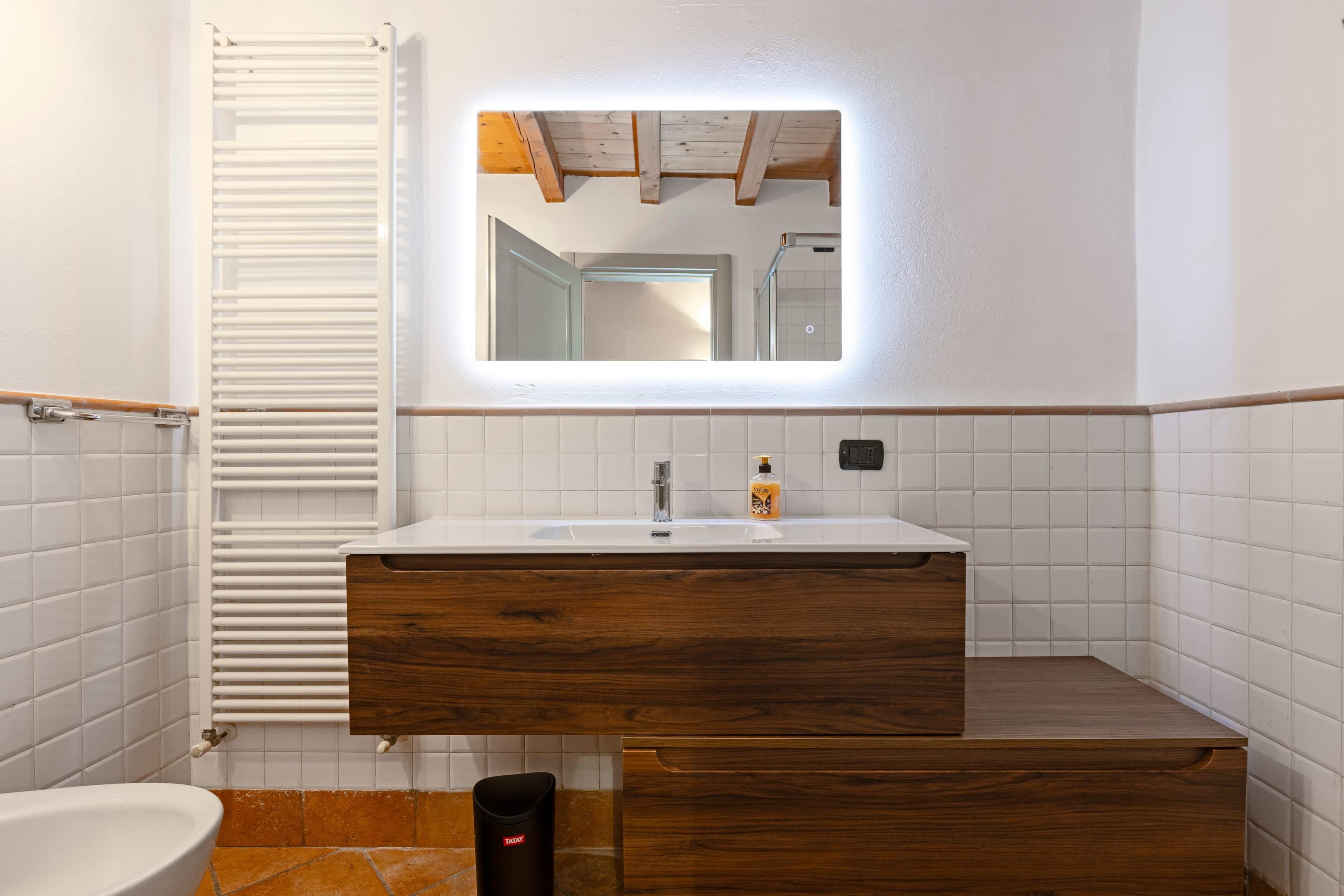 A bathroom with a white sink, wooden vanity, and a mirror with integrated lighting.