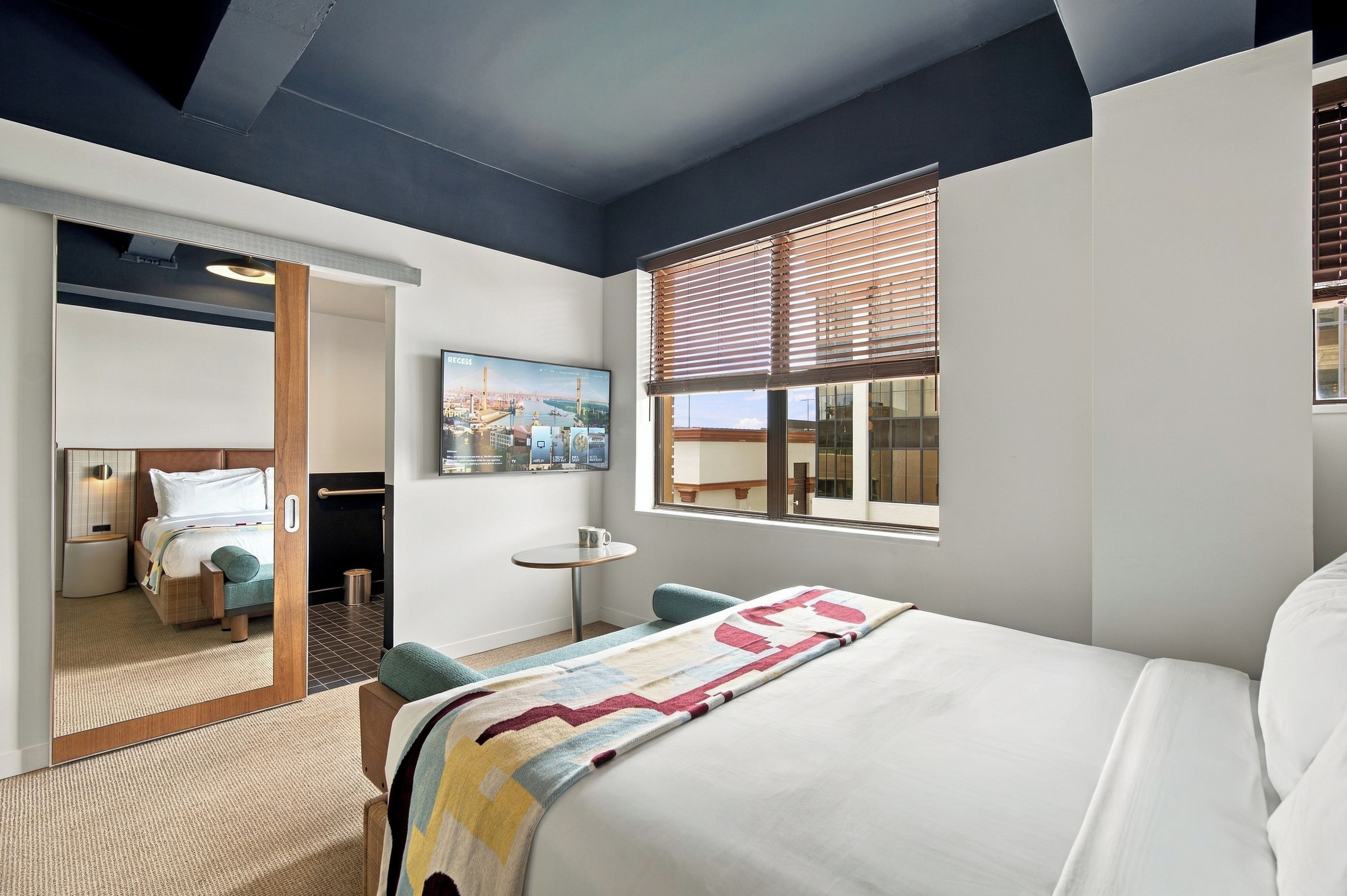 A hotel room with a bed, a small table, a window with blinds, a painting on the wall, and a mirror reflecting the room.