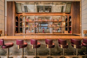 Una zona de bar con varias estanterías llenas de botellas, una barra central y varios taburetes de bar.