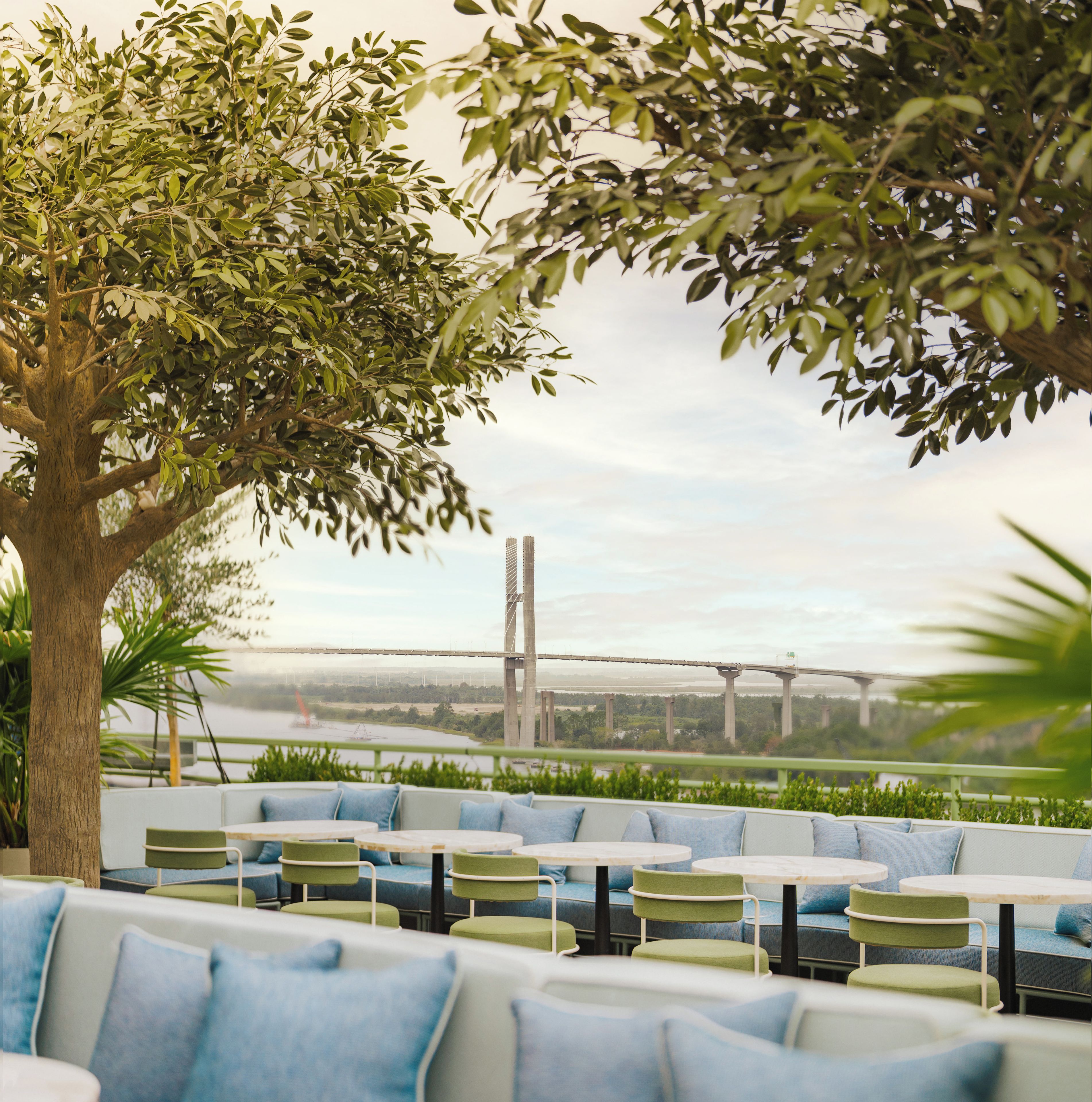 An outdoor seating area with tables and chairs, a large tree, and a view of a bridge and river.