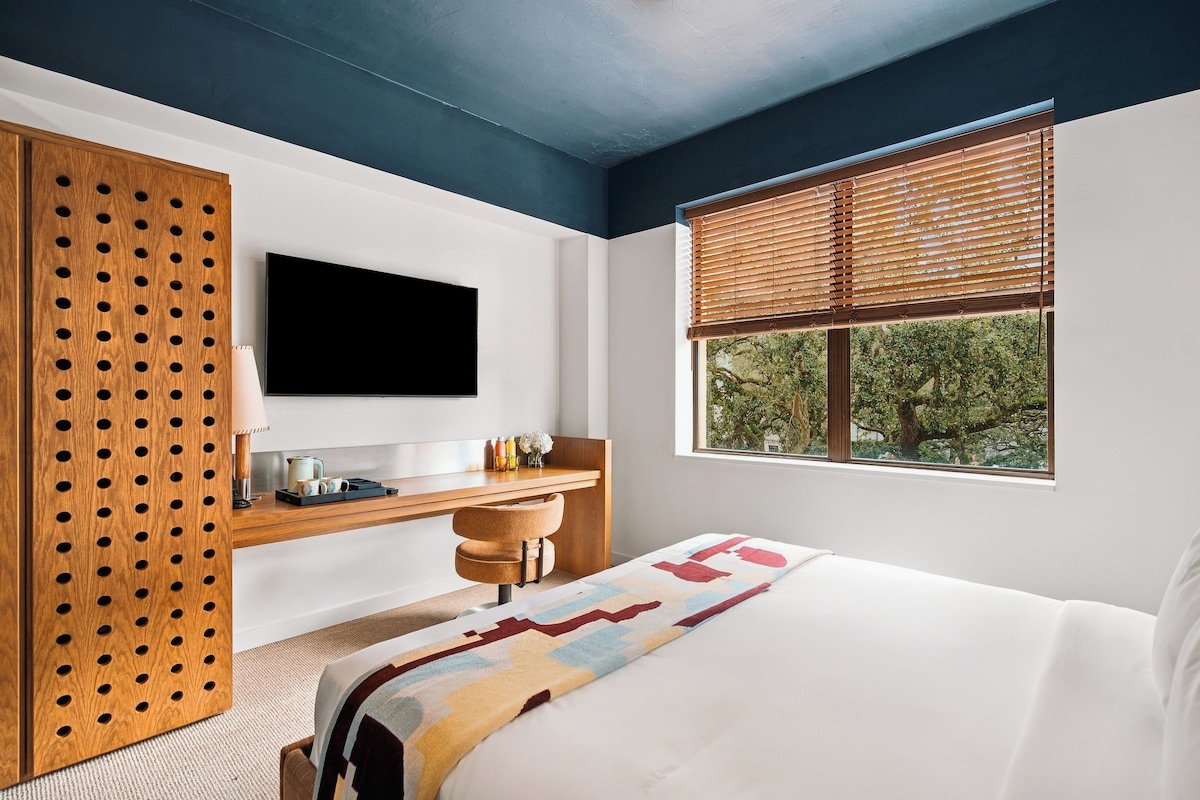 A bedroom with a bed, a desk, a television, and a window with blinds.