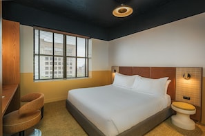 A hotel room with a bed, a desk, a chair, a toilet, and a window with blinds. - Recess Hotel and Club (Savannah)