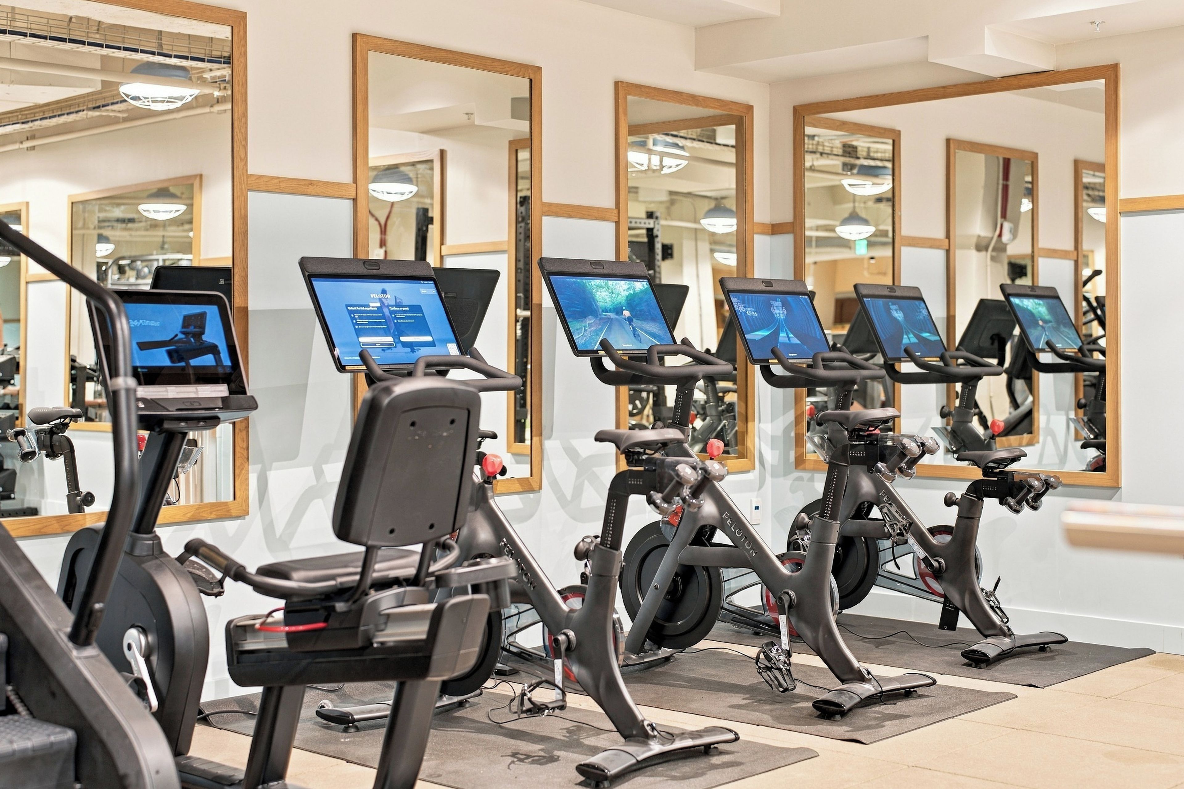 A row of stationary bikes with attached screens, likely for fitness or entertainment purposes, in a well-lit indoor space with mirrors on the walls.