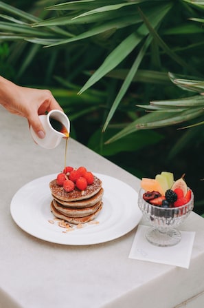 A hand pouring syrup over a stack of pancakes on a white plate, with a bowl of fruit next to it.