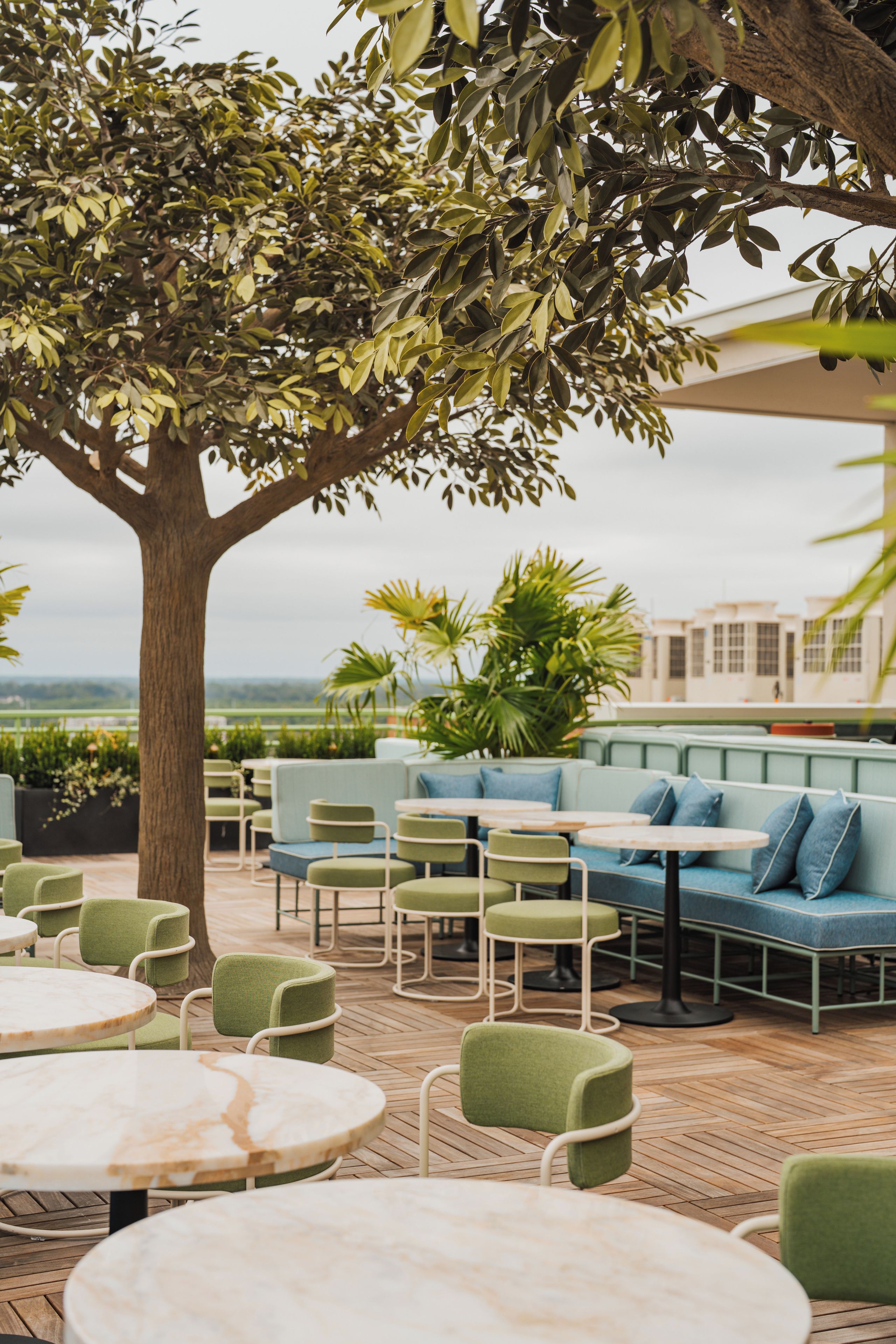 An outdoor seating area with round tables and green chairs, surrounded by a tree and potted plants.