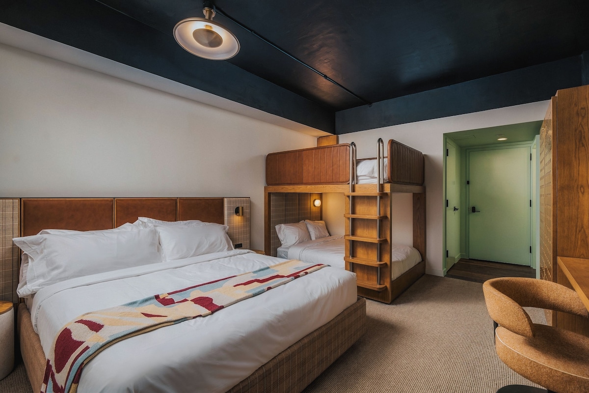 A hotel room with a large bed, a wooden bunk bed, a chair, and a door.