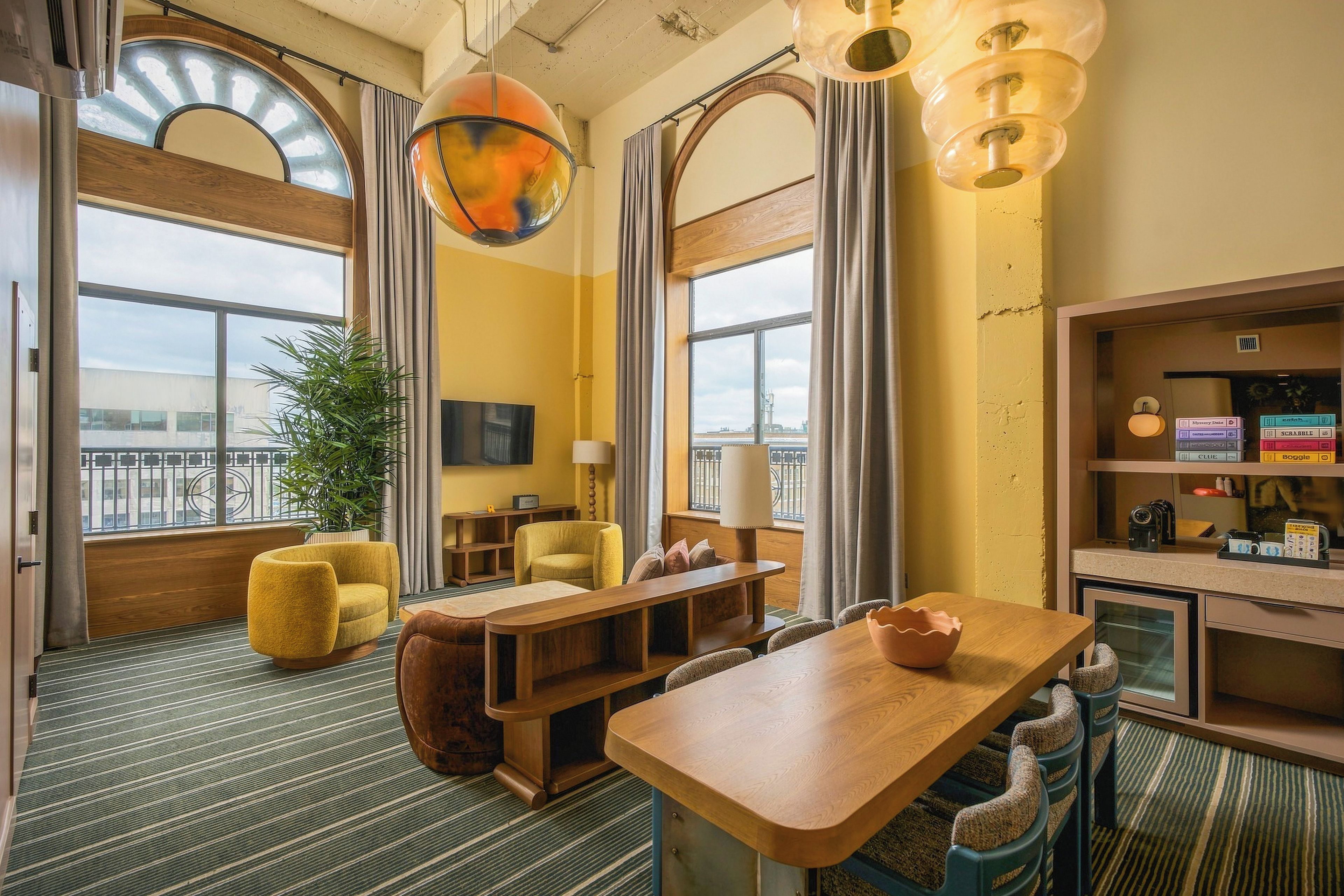 A hotel room with a wooden dining table, two yellow armchairs, a large potted plant, and a bookshelf with board games.