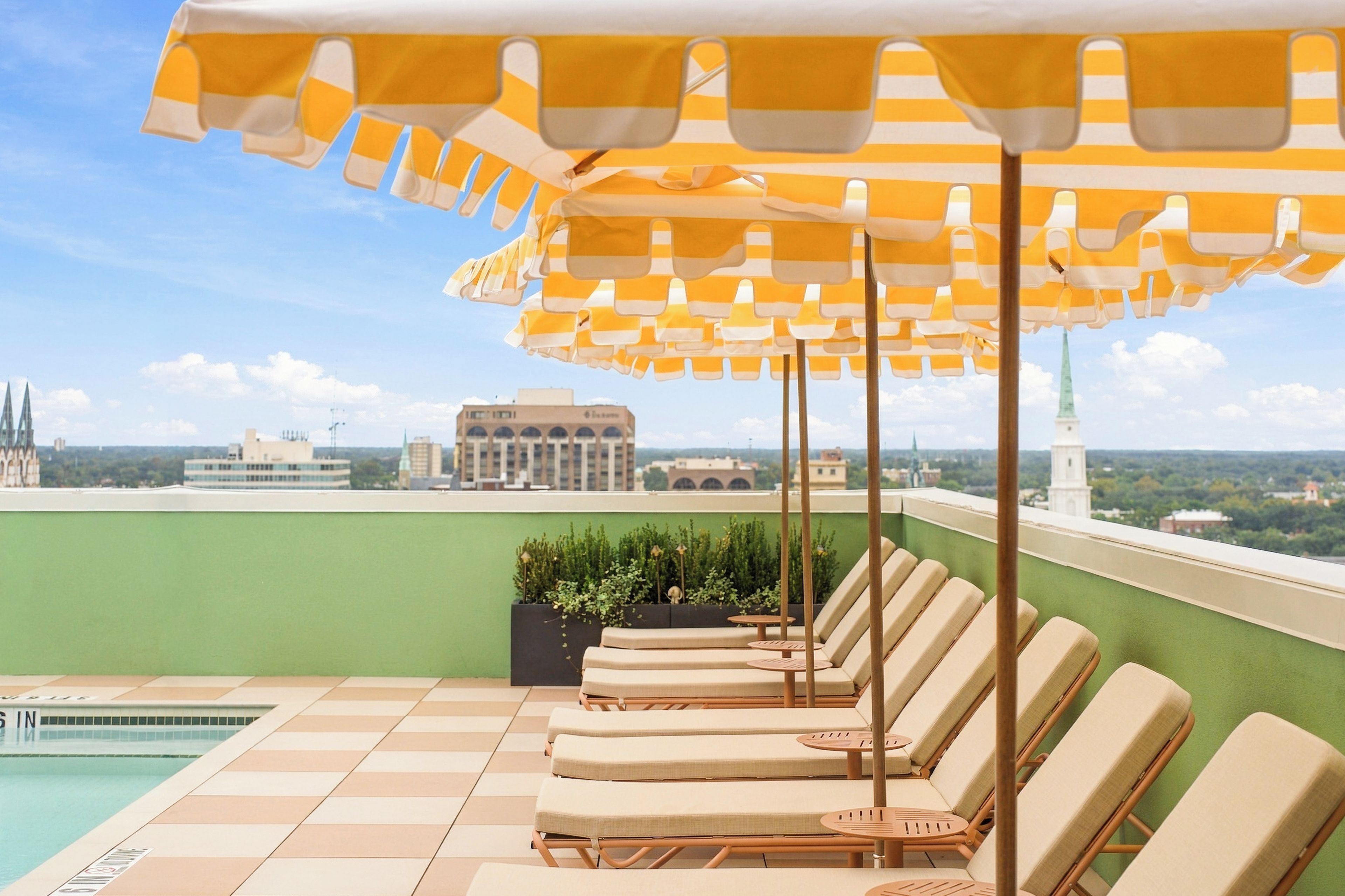 A rooftop pool area with lounge chairs and striped umbrellas, overlooking a cityscape.