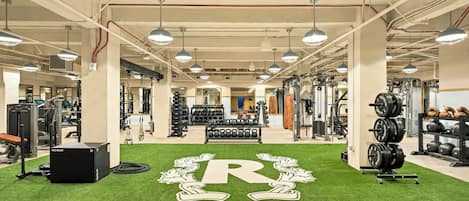 A gym with various exercise equipment, artificial turf with a logo, and overhead lighting.