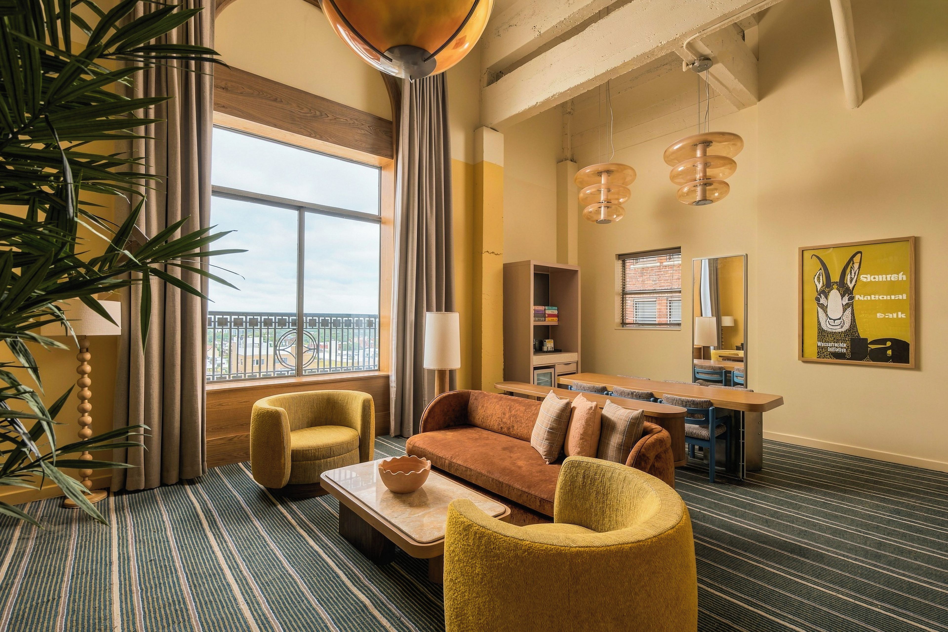 Une chambre d’hôtel comprenant un canapé, des fauteuils, une table basse, une table à manger avec des chaises, une bibliothèque, une grande fenêtre avec vue et un luminaire suspendu.