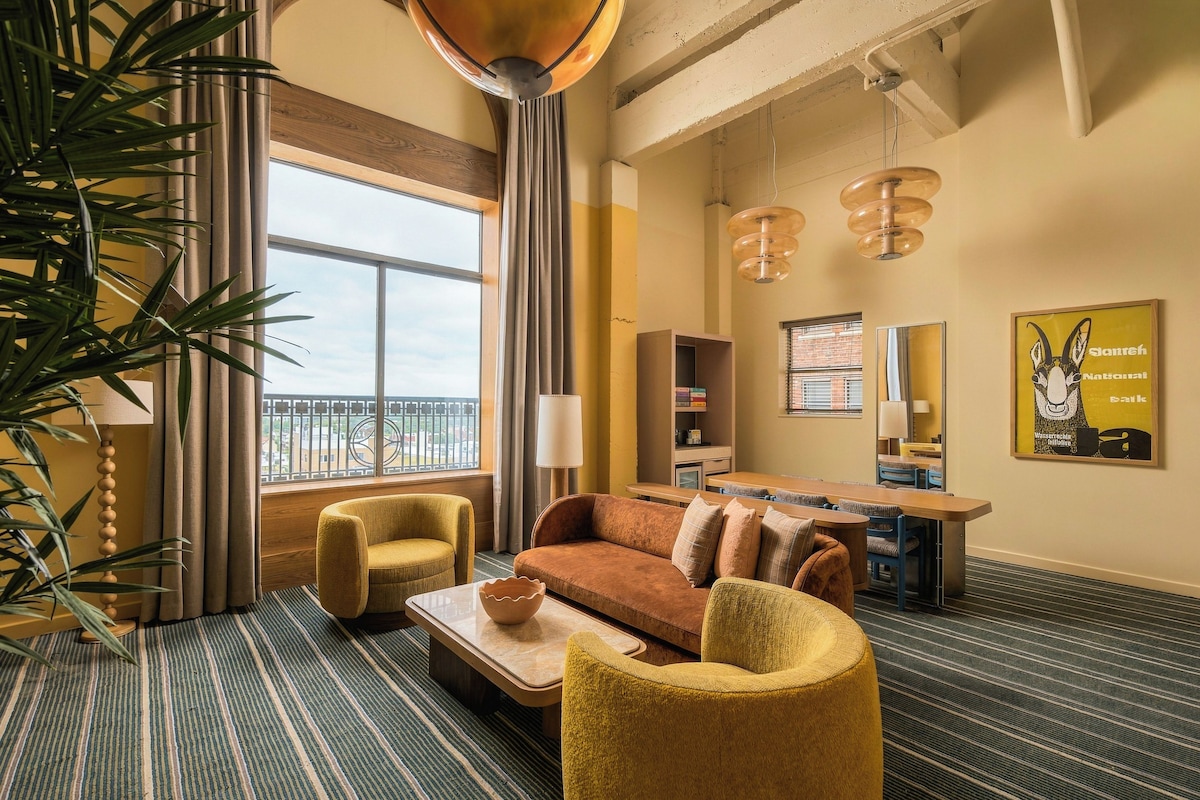 A hotel room with a sofa, armchairs, a coffee table, a dining table with chairs, a bookshelf, a large window with a view, and a hanging light fixture.