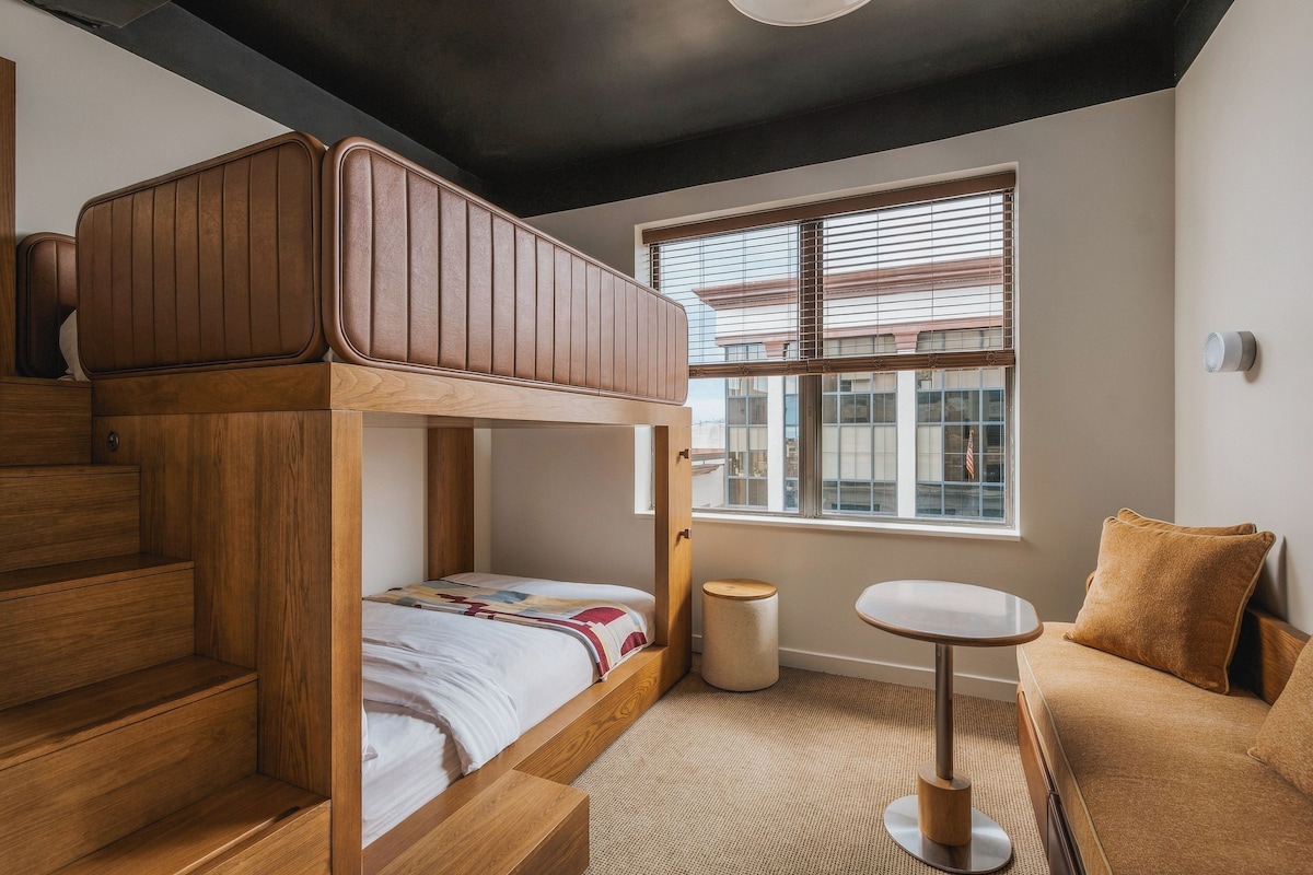 A bedroom with a bunk bed, a small round table, a chair, and a window with blinds.