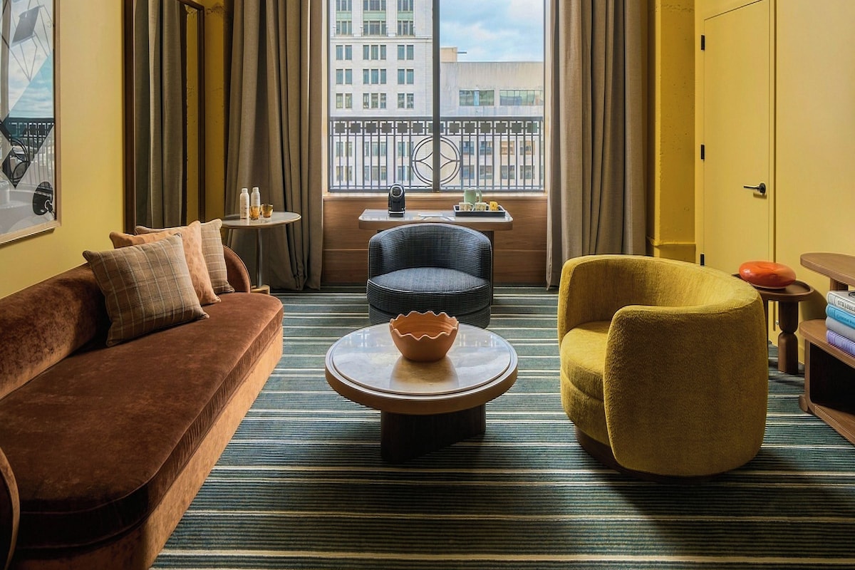 A hotel room with a large window, a brown sofa, a yellow armchair, a round coffee table, and a colorful pendant light.