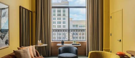 A hotel room with a large window, a brown sofa, a yellow armchair, a round coffee table, and a colorful pendant light.