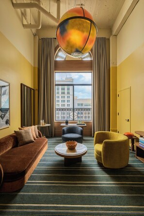 A hotel room with a large window, a brown sofa, a yellow armchair, a round coffee table, and a colorful pendant light. - Recess Hotel and Club (Savannah)