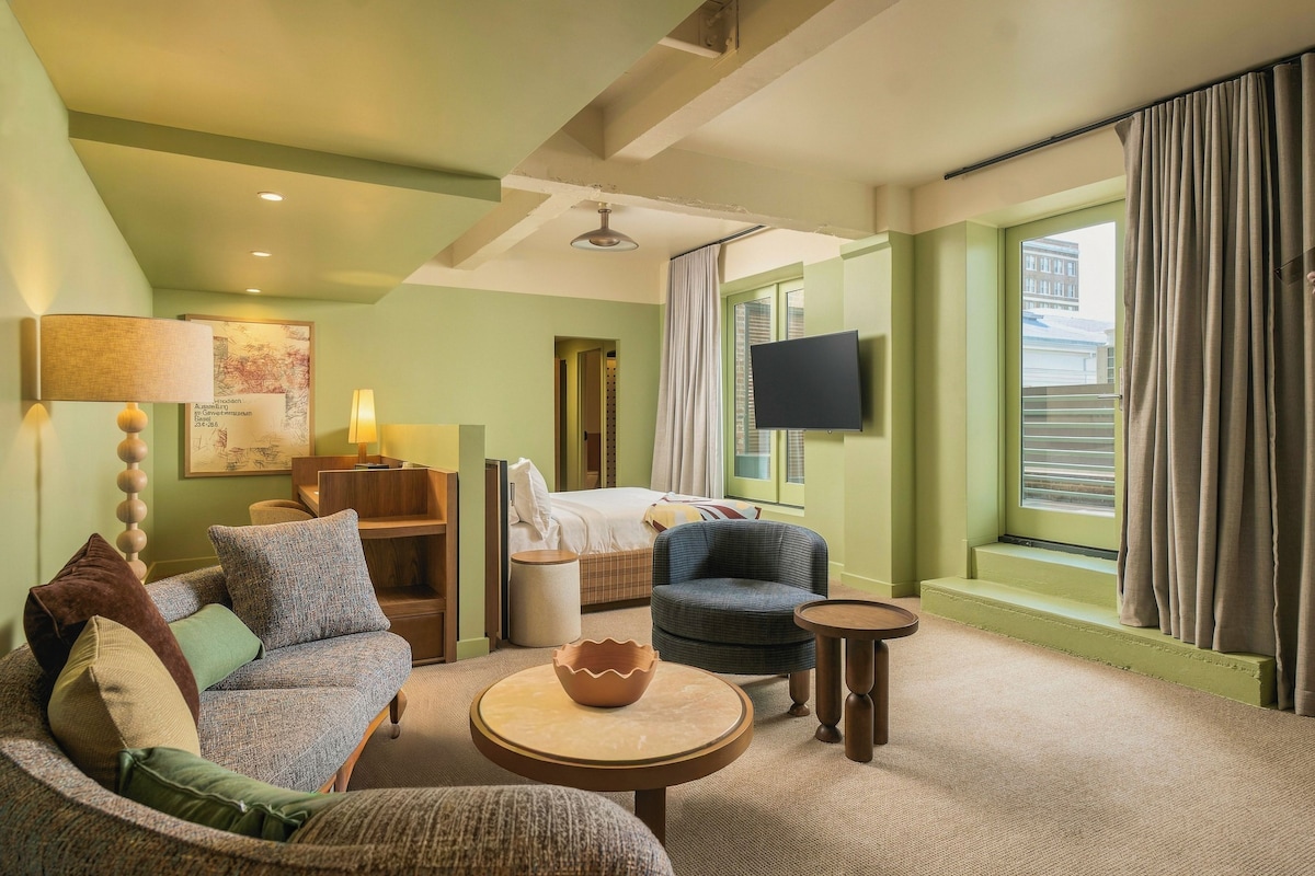 A hotel room with a bed, a sofa, a chair, a small table, a lamp, a television, and a window with curtains.