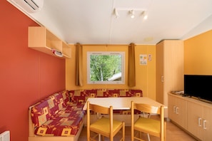 Ein Zimmer mit einer Couch, einem Tisch, Stühlen, einem Fernseher und einem Fenster mit Blick auf Bäume.