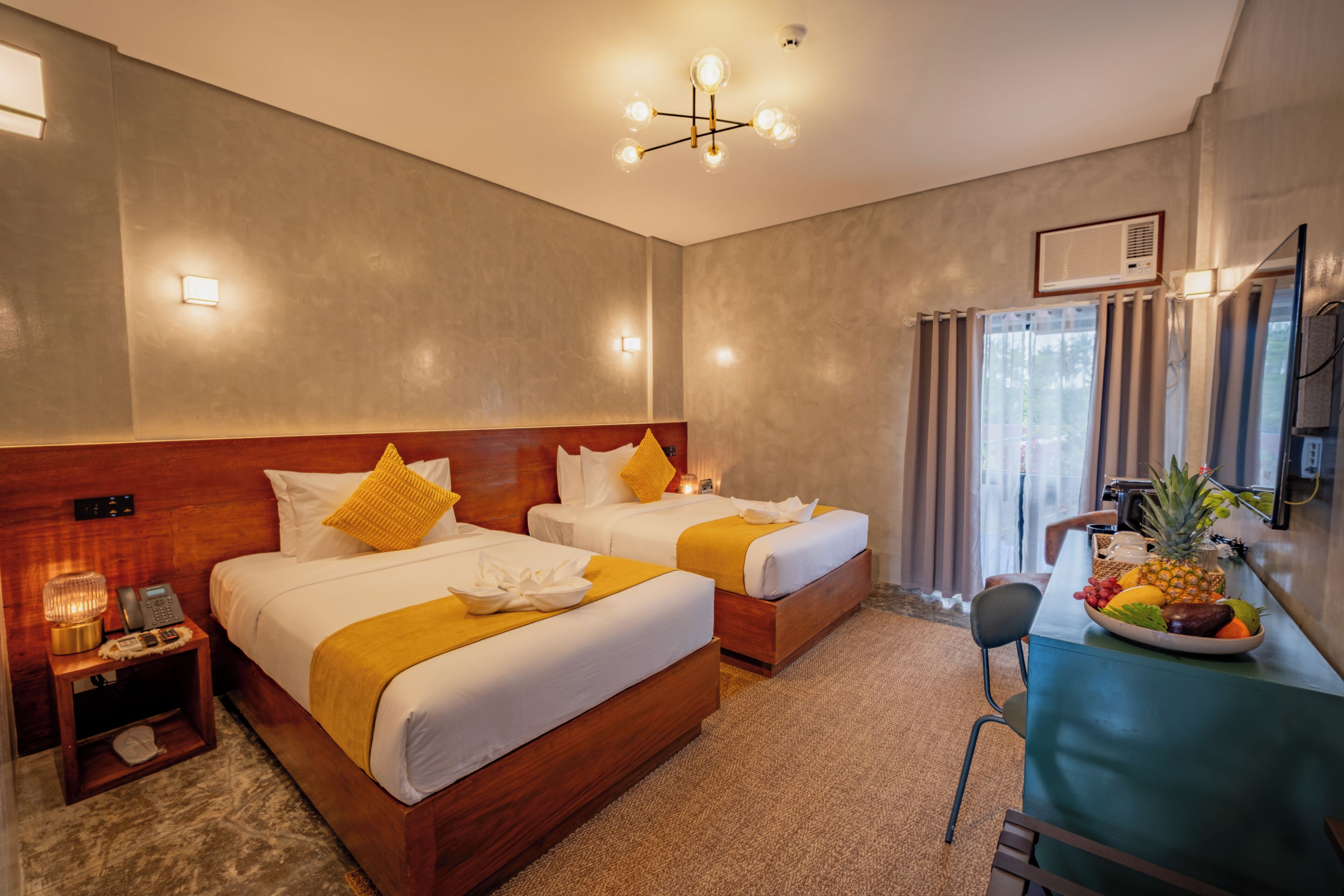 A hotel room with two beds, a desk with a fruit bowl, a chair, a small table with a lamp, and a ceiling light fixture.