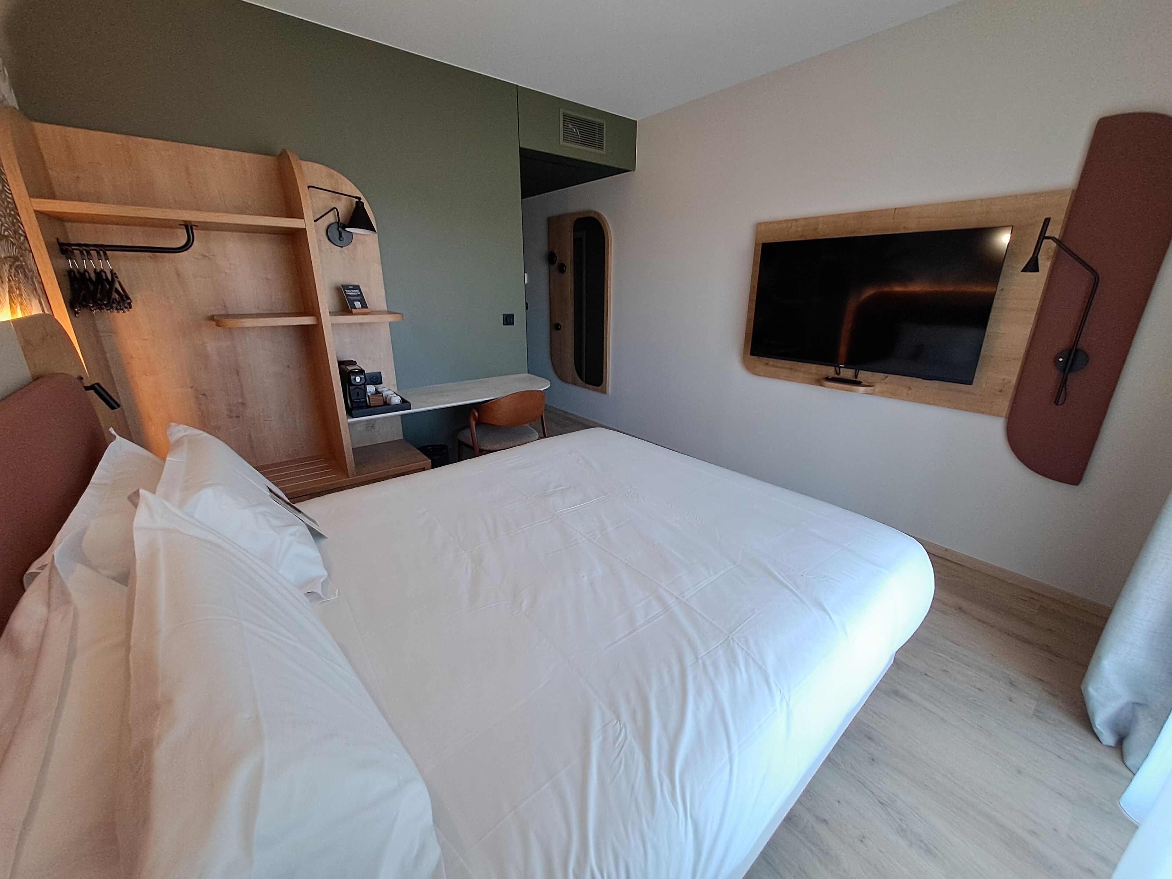 A hotel room with a bed, a desk, a chair, a mirror, and a television.