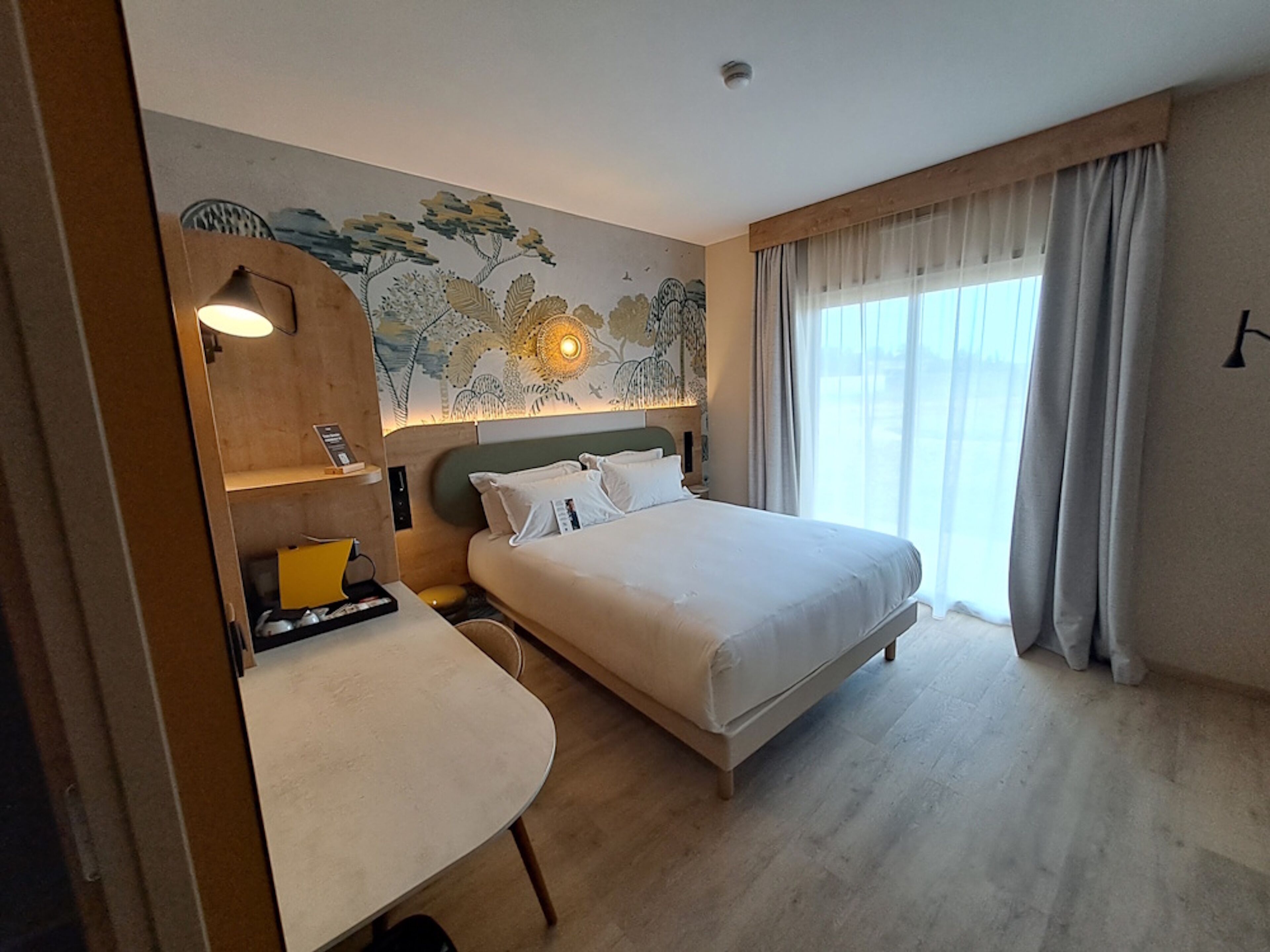 Une chambre d’hôtel avec un lit, un bureau, une chaise, une lampe et une grande fenêtre avec des rideaux.