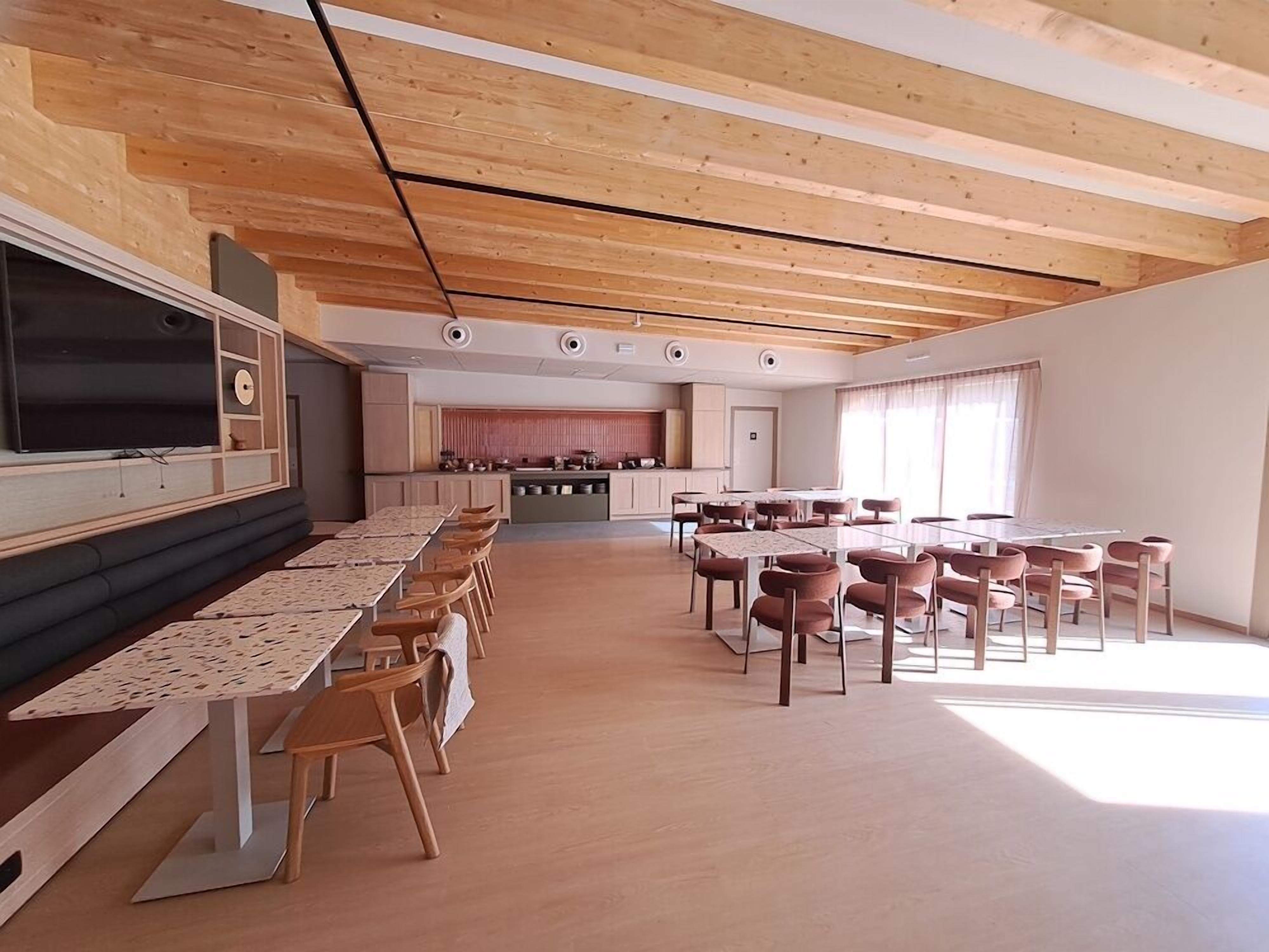 Um quarto espaçoso com uma mesa comprida e cadeiras, uma área de cozinha ao fundo, e teto de madeira.