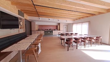 Um quarto espaçoso com uma mesa comprida e cadeiras, uma área de cozinha ao fundo, e teto de madeira.