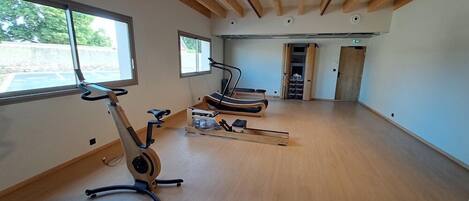 A spacious room with wooden beams on the ceiling, exercise bikes, and a large window providing natural light.