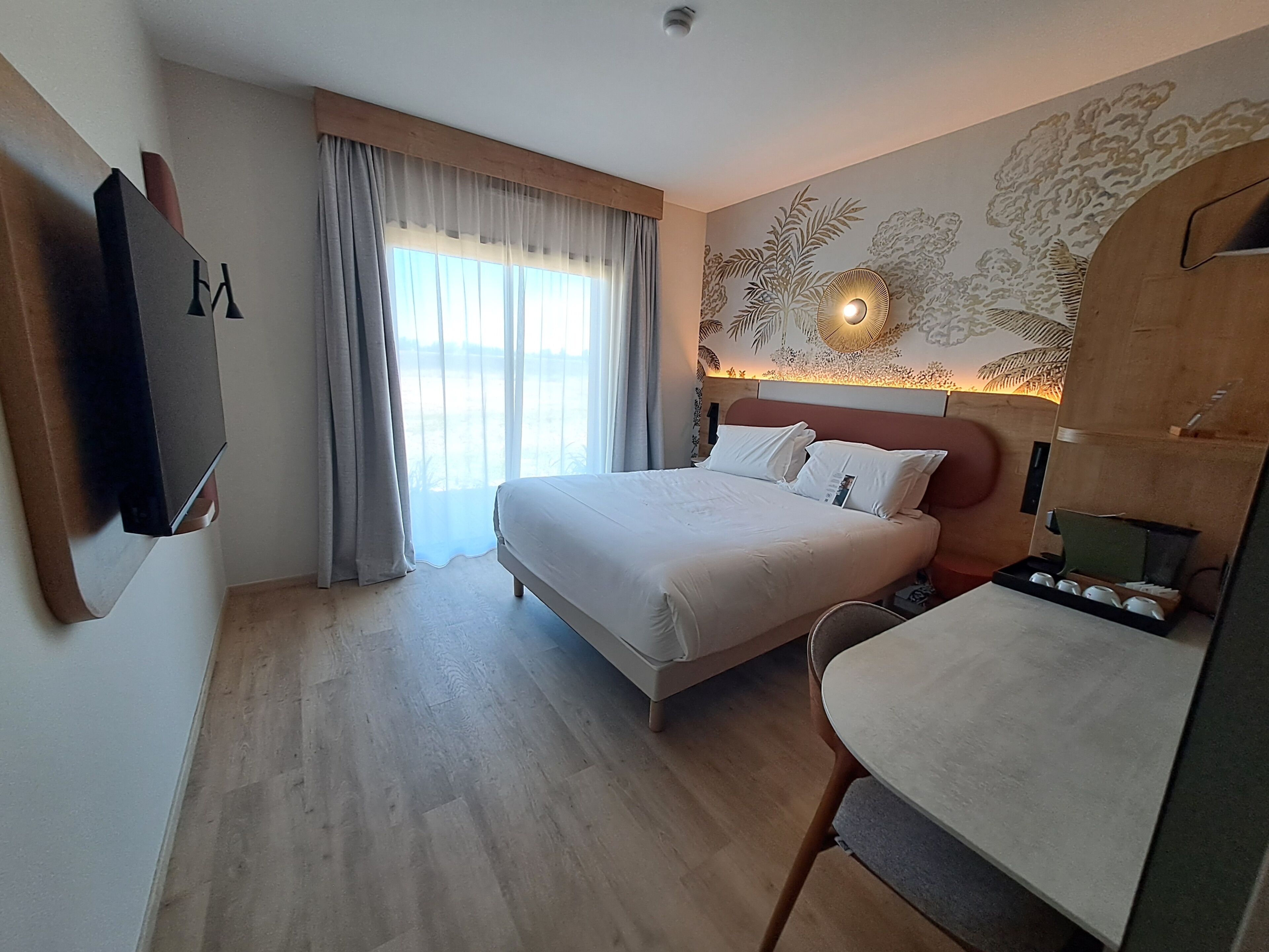 A hotel room with a bed, a desk, a chair, a flat-screen TV, and a large window with curtains.