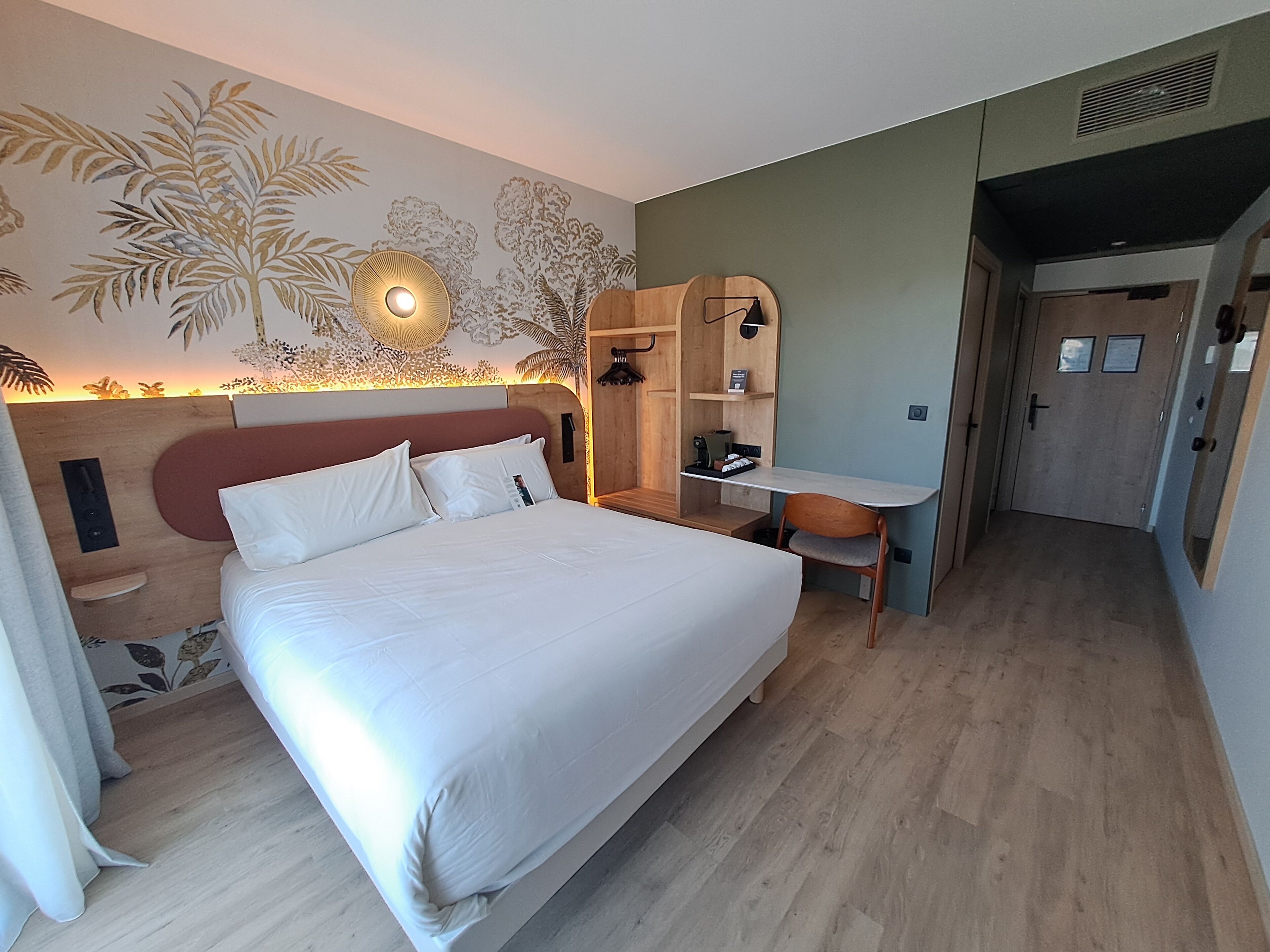 A hotel room with a bed, a desk, a chair, a mirror, and a wall with a tropical mural.