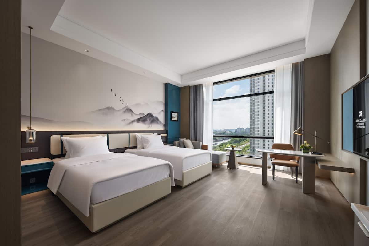 Una habitación de hotel moderna con dos camas, un escritorio, una silla, un ventanal con vistas a la ciudad y un televisor montado en la pared.