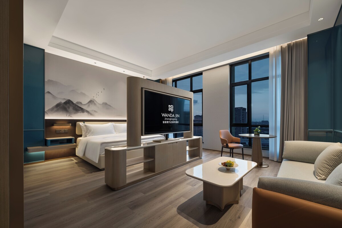 A modern hotel room with a bed, a TV, a sofa, a coffee table, and a city view.
