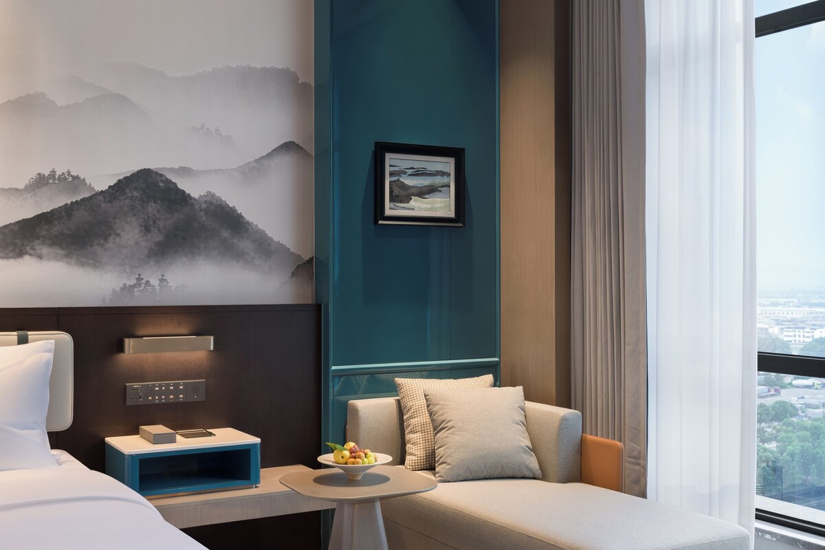 A modern hotel room with a bed, a sofa, a coffee table, and a large window with a view.