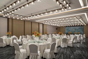 Un salón de banquetes con mesas redondas, sillas blancas e iluminación colgante.