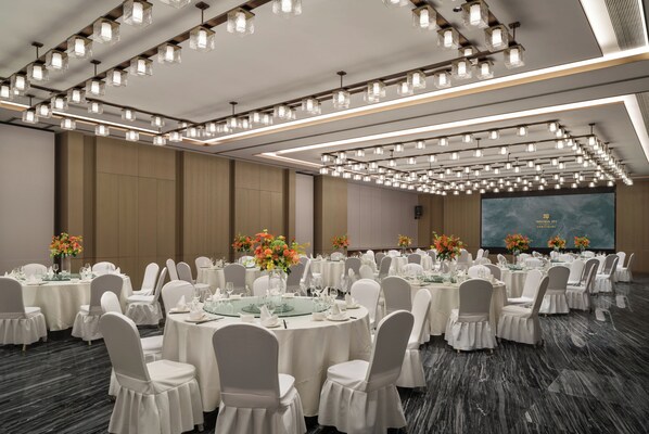 Un salón de banquetes con mesas redondas, sillas blancas e iluminación colgante.