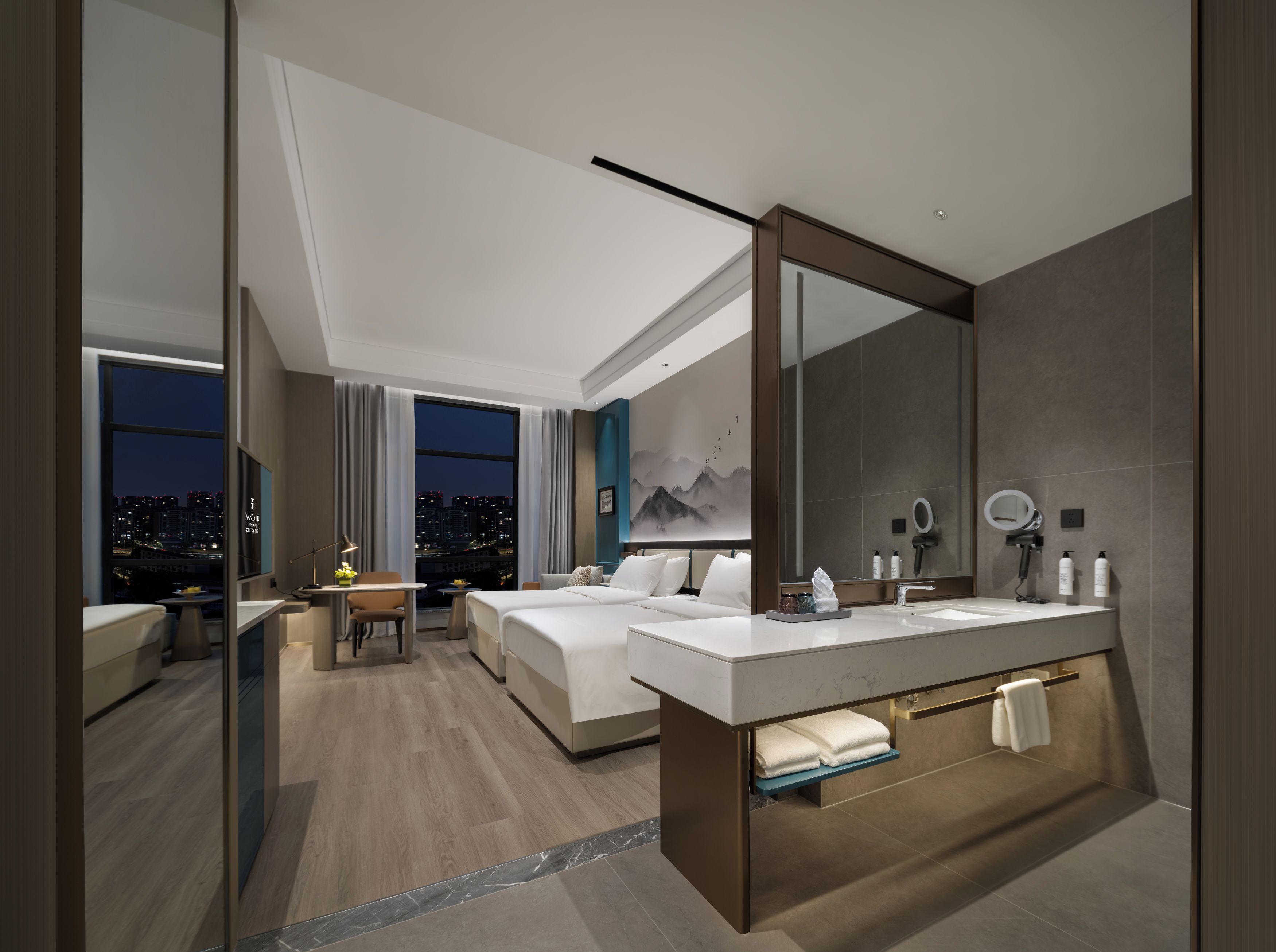 A modern hotel room with a large bed, a desk, a bathroom with a vanity, and a city view.
