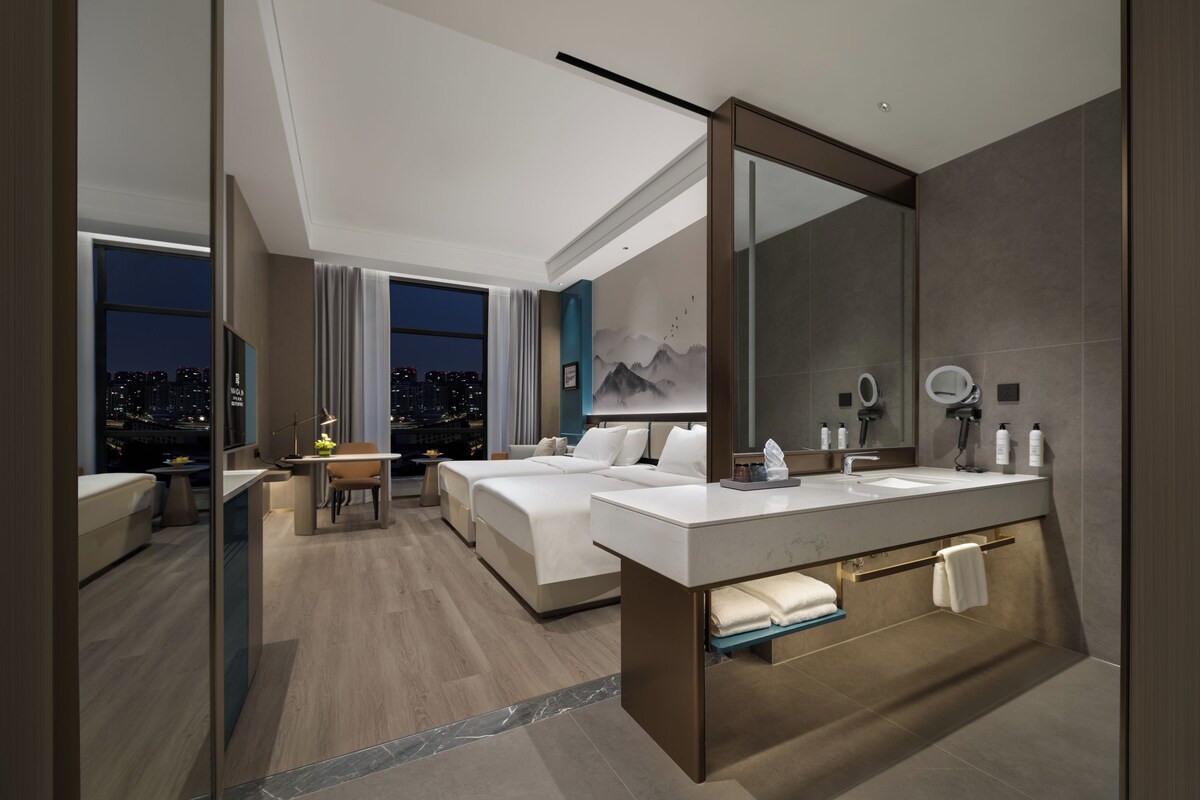 A modern hotel room with a large bed, a desk, a bathroom with a vanity, and a city view.