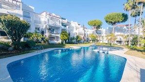 A swimming pool surrounded by modern buildings with balconies, lush greenery, and palm trees. - Apartment in Cancelada Near the Beach (Estepona)