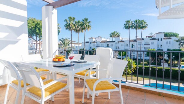 A terrace with white furniture and yellow cushions, a table with plates and a glass of red wine, and a view of white buildings and palm trees. - Apartment in Cancelada Near the Beach (Estepona)