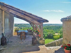 A stone patio with a wooden pergola, outdoor seating, and a view of the countryside.