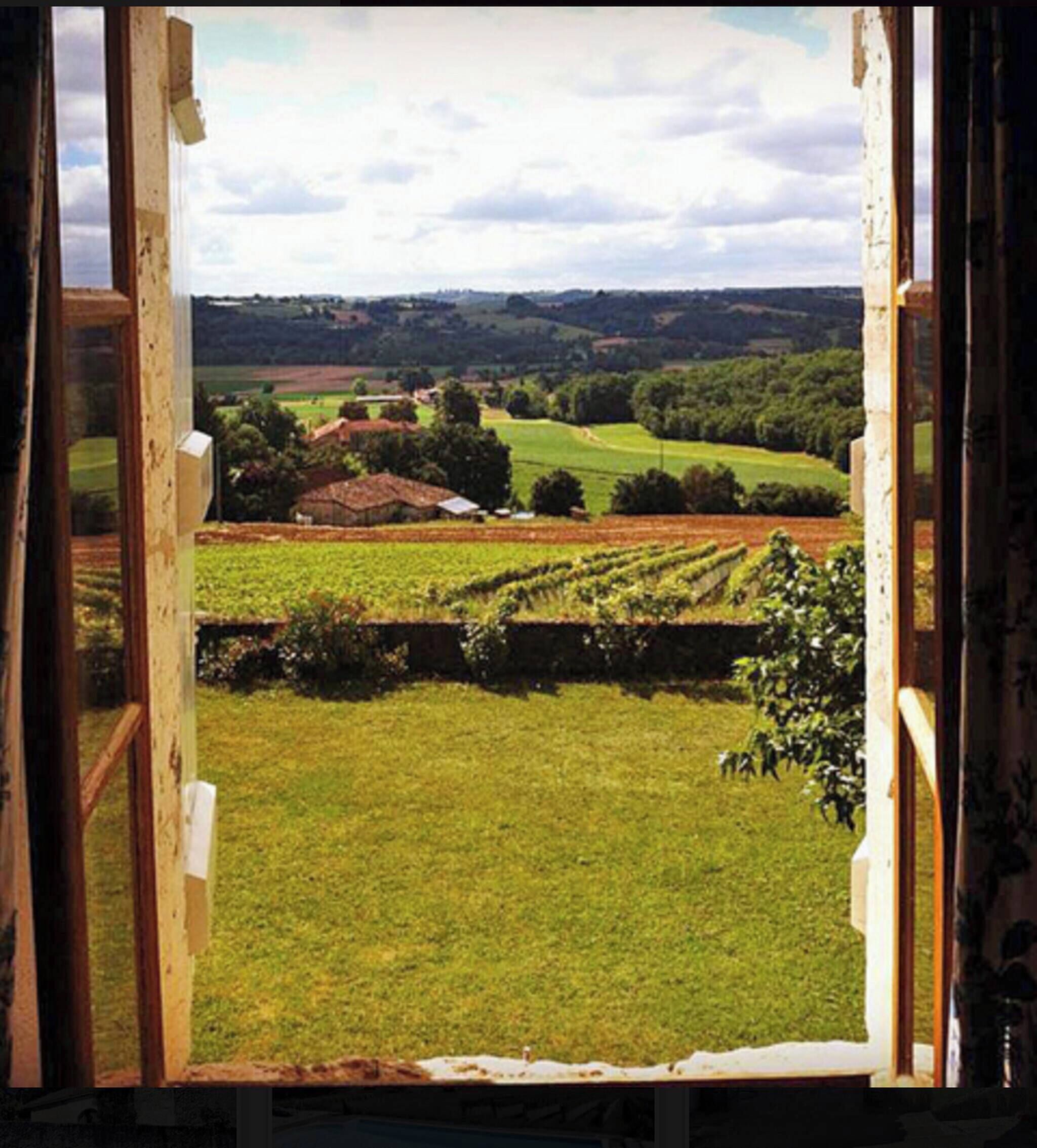 Une vue à travers une fenêtre sur un paysage rural, avec des champs, une petite maison et des collines au loin.