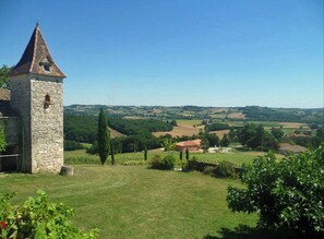 A stone tower with a conical roof, surrounded by a grassy field and vineyards, overlooking rolling hills.