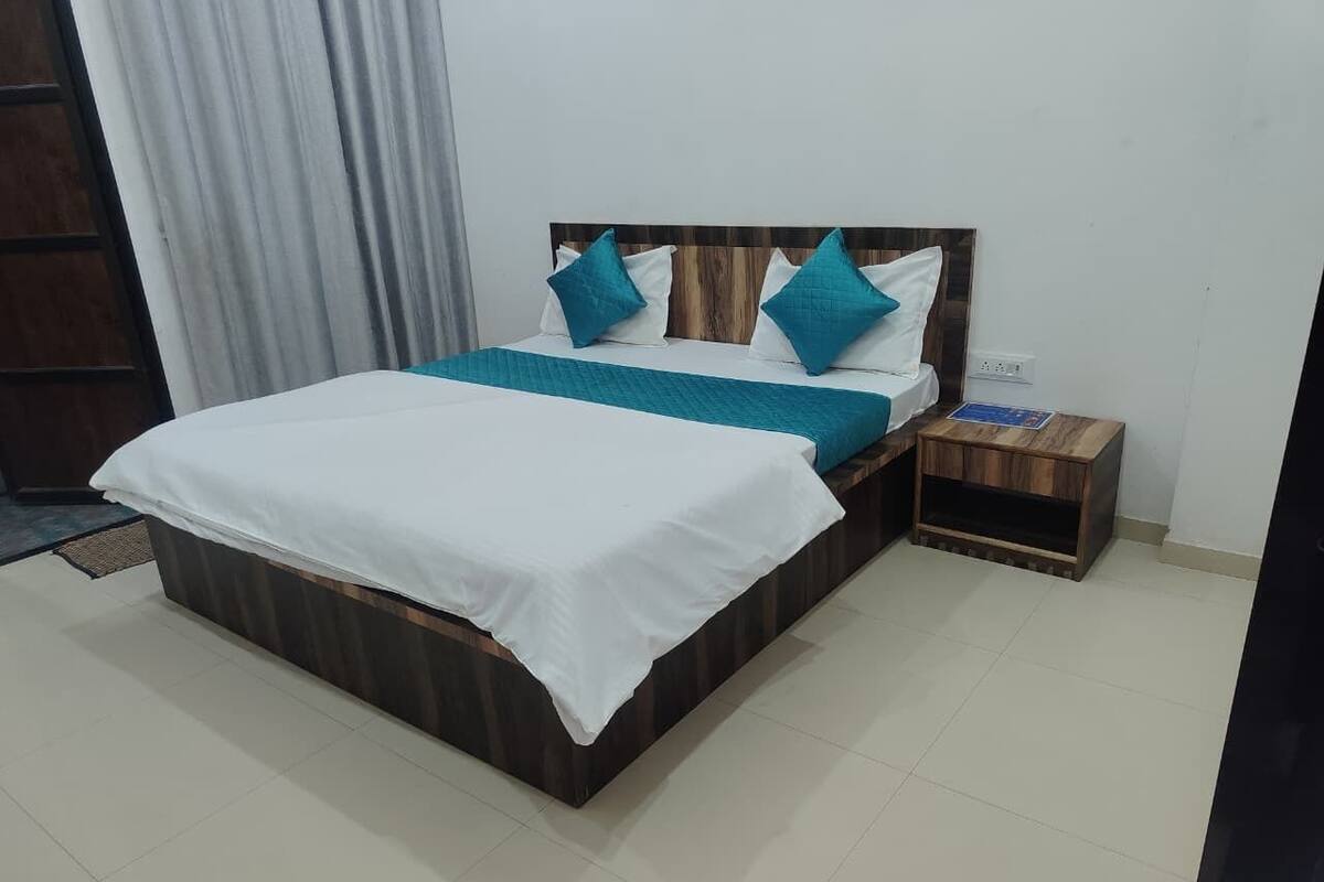 Un lit avec une tête de lit en bois, une literie blanche et des coussins turquoise. Il y a une petite table de chevet en bois avec une lampe et une prise de courant.
