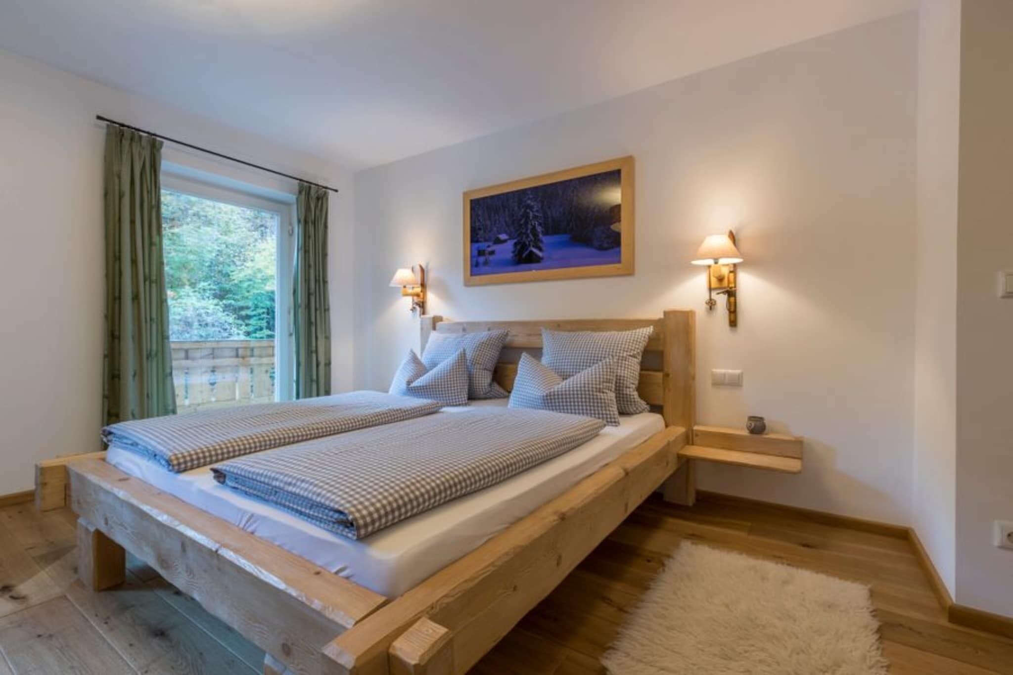 Ein Schlafzimmer mit einem hölzernen Bett, zwei Nachttischen, einem Beistelltisch, einem Fenster mit Vorhängen und einem Bild an der Wand.