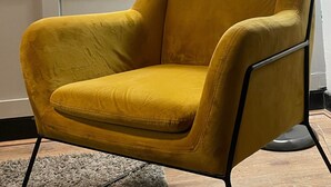 Un sillón amarillo mostaza con estructura metálica negra, ubicado frente a una pared con obras de arte enmarcadas y una lámpara de pie.