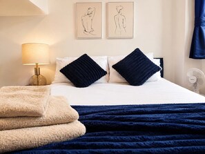 Ein ordentlich bezogenes Bett mit zwei Kissen, einer Nachttischlampe, einem Ventilator und Bildern an der Wand.
