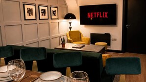 Un comedor con una mesa dispuesta para cuatro, un sillón amarillo, una pantalla de televisión negra mostrando 'NETFLIX' y obras de arte enmarcadas en la pared.