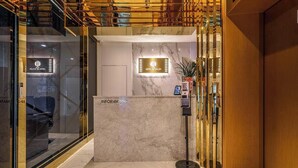 Zona de recepción de hotel con un mostrador de mármol, un quiosco digital informativo y una pared de vidrio con vista al interior.