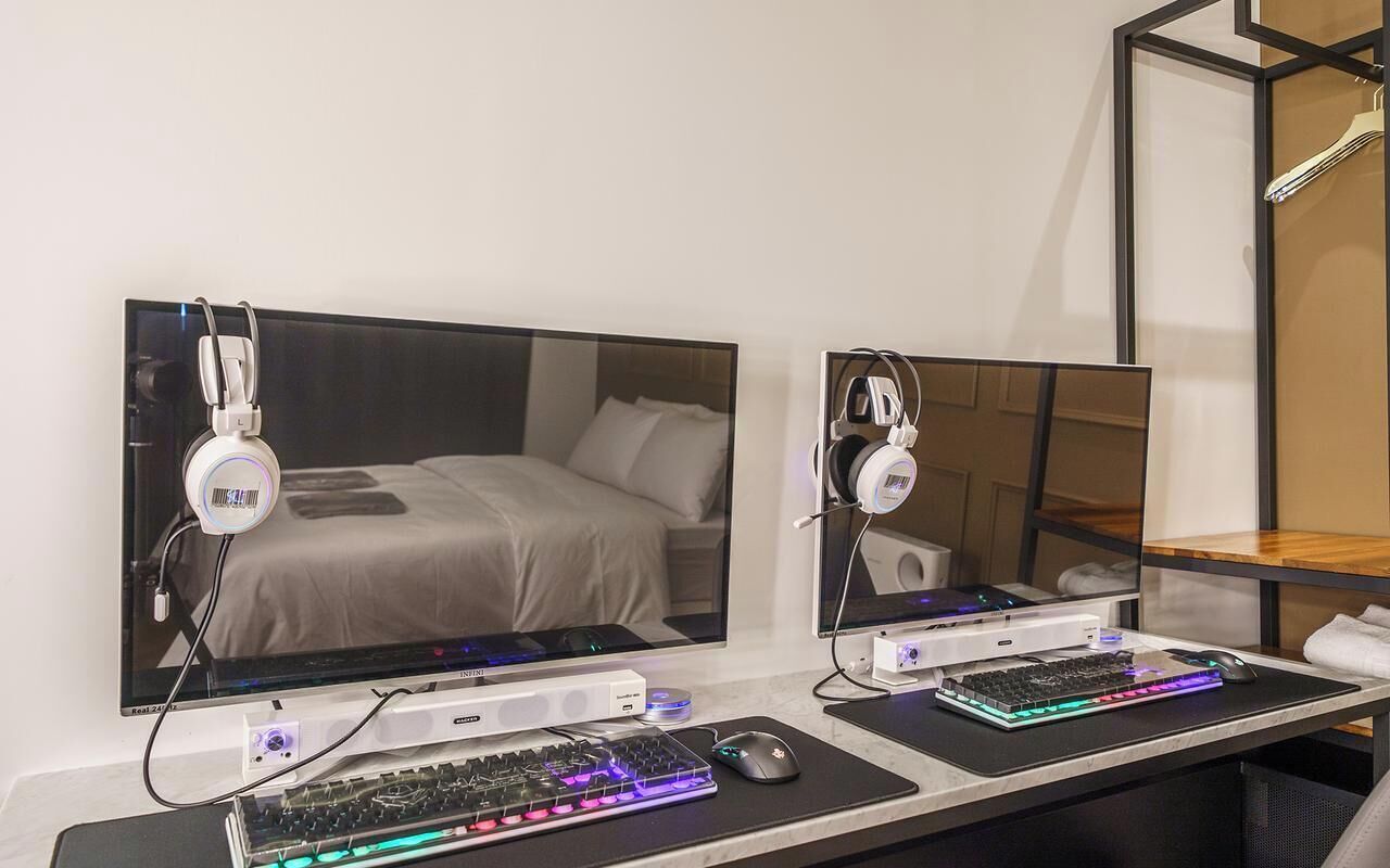 A modern hotel room with a large bed, two monitors, a gaming setup, and a mirror.