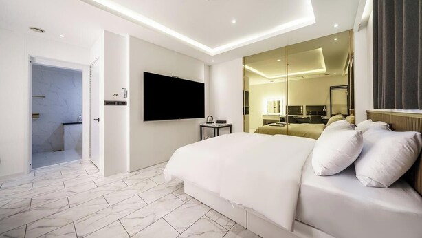 Una habitación de hotel moderna con una cama grande, un televisor de pantalla plana montado en la pared, una mesita auxiliar pequeña y un baño visible a través de una puerta abierta.